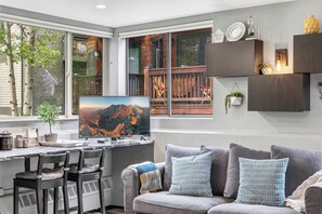 Ein modernes Wohnzimmer mit großem Fernseher, einer Couch mit Kissen, einem Schreibtisch mit Stühlen und einem Fenster mit Bergblick.