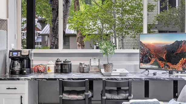 A kitchen with a coffee maker, sugar, coffee containers, a potted plant, and a TV displaying a mountainous landscape. - Cozy Mountain Nook-Tomboy Studio #133 (Telluride)