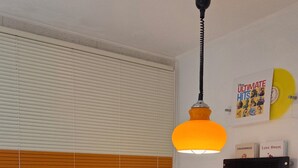 Ein Zimmer mit einem weißen Tisch, schwarzen Stühlen, einem grünen Hocker, einer hängenden orangefarbenen Pendelleuchte und einem Fenster mit Jalousien.