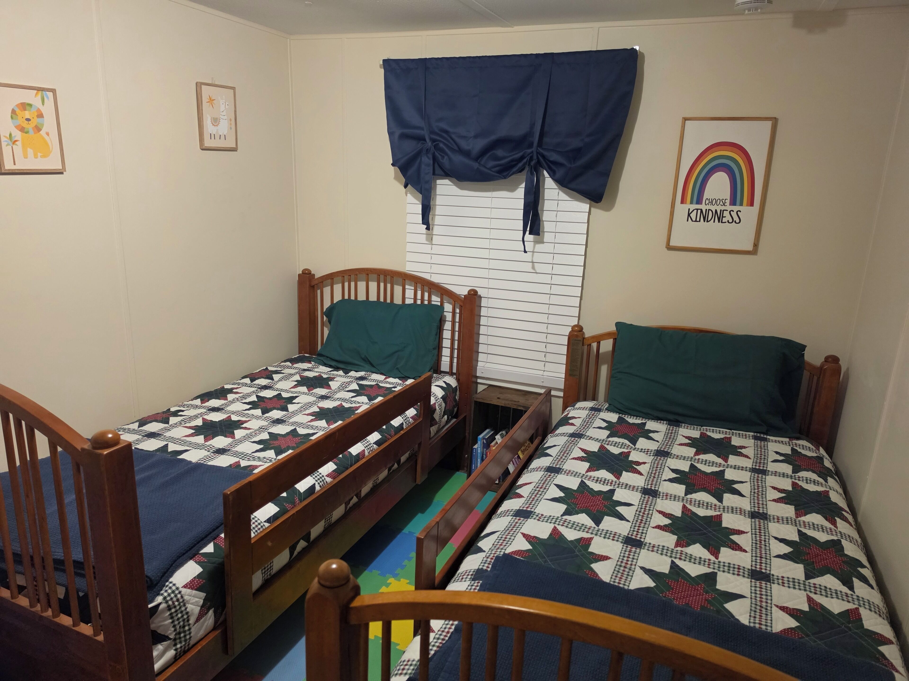 Une chambre à coucher avec deux lits en bois, chacun recouvert d’une courtepointe matelassée et agrémenté d’un oreiller. Il y a une fenêtre avec des stores et un rideau bleu.