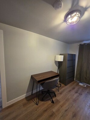 Et værelse med skrivebord i træ, stol, gulvlampe og et mørkt skab.