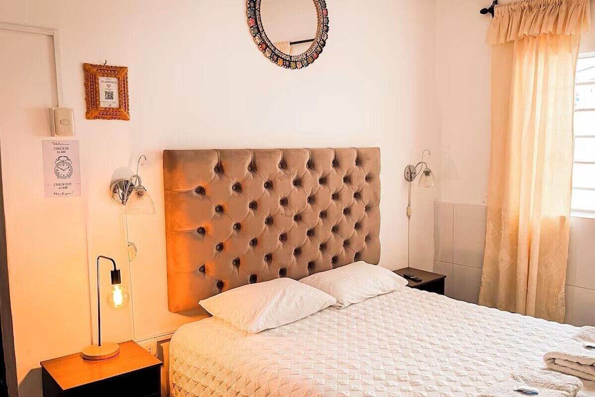 A bedroom with a tufted headboard, a bedside lamp, a framed picture, and a window with curtains.