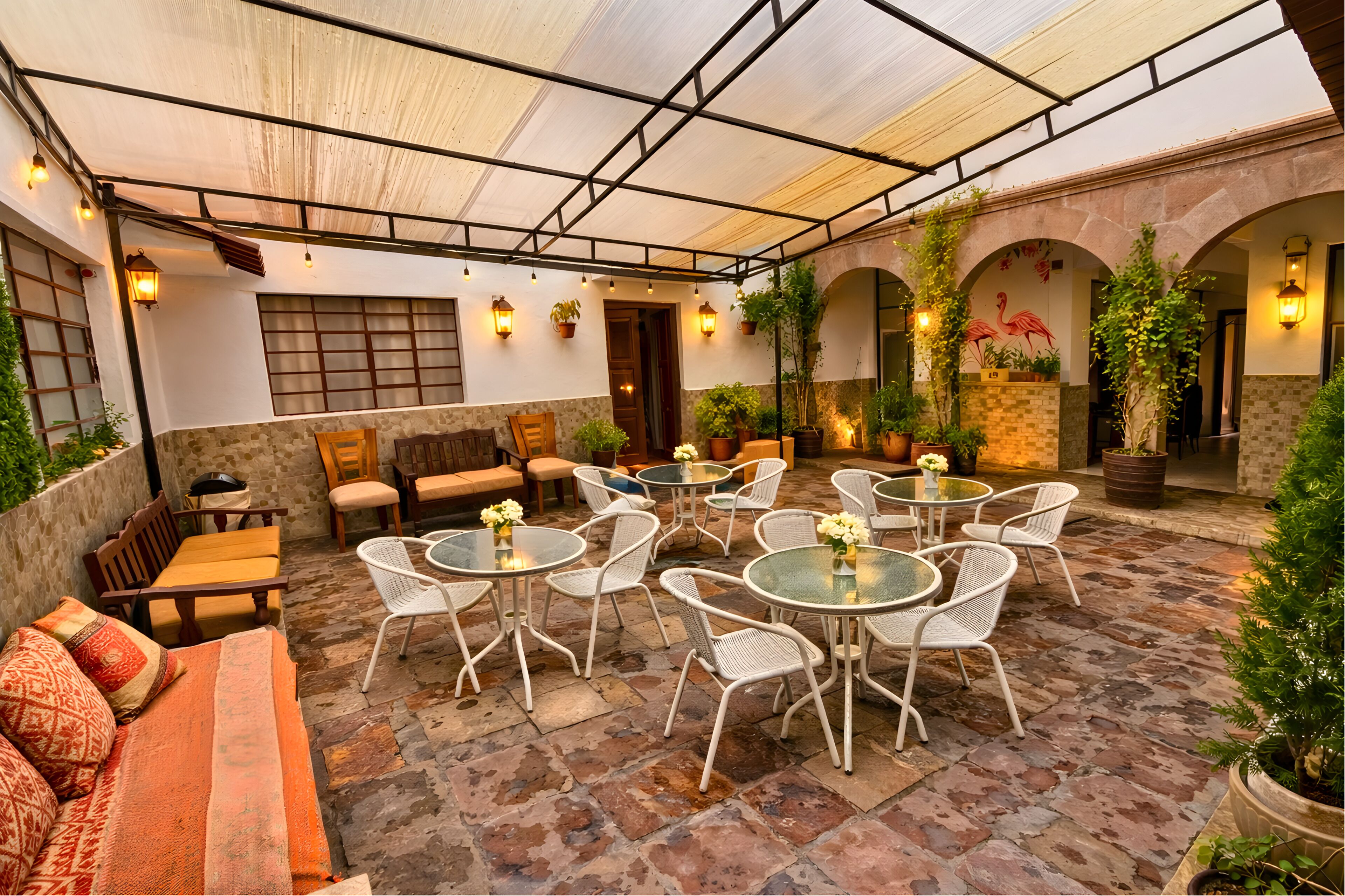 A covered outdoor seating area with tables and chairs, potted plants, and a stone floor.