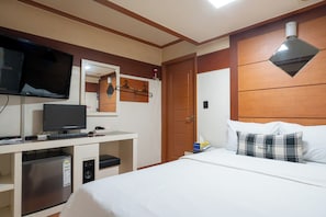 Habitación de hotel con cama, televisión, escritorio con computadora, refrigerador y cabecera de madera.