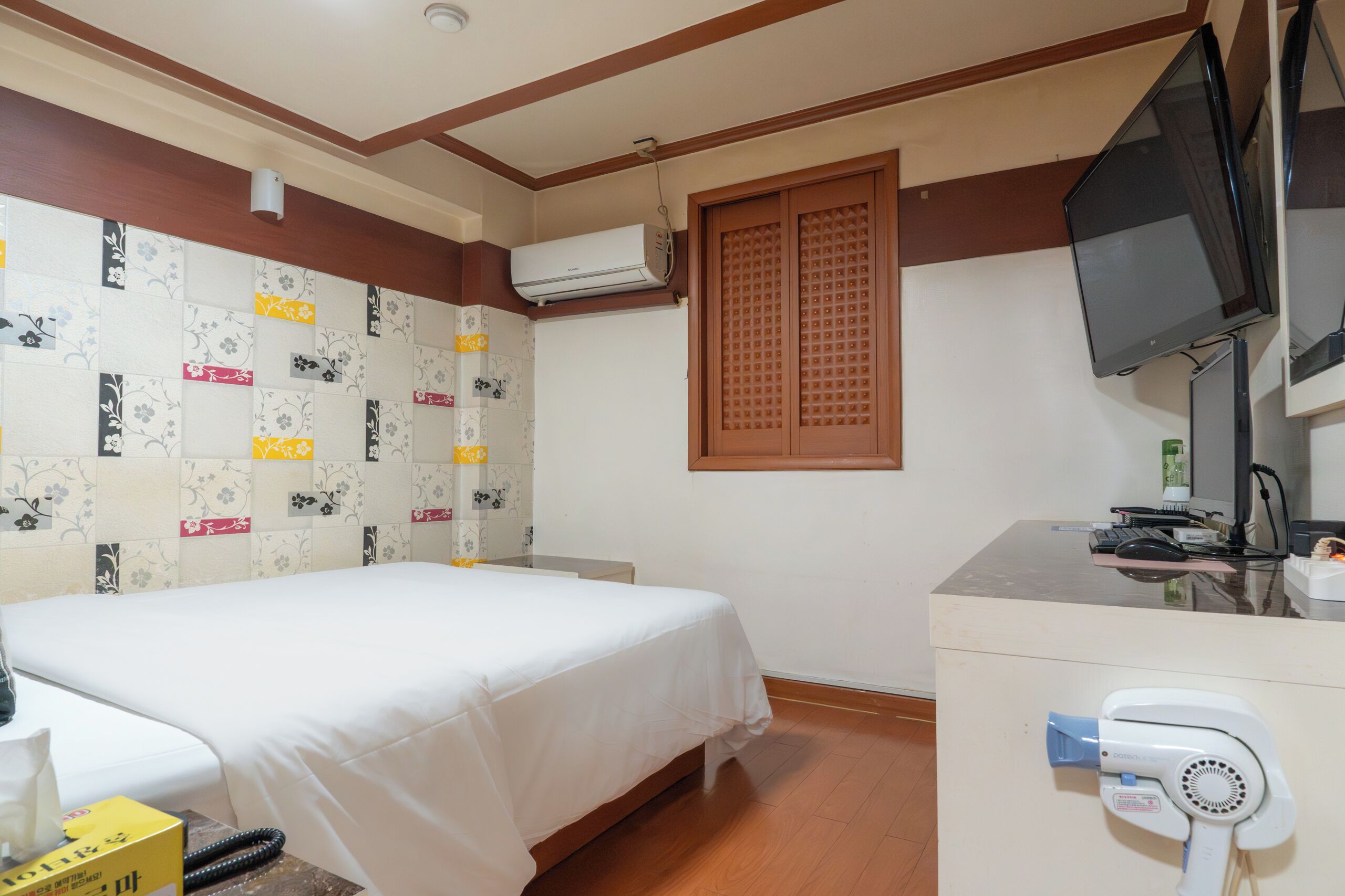 Habitación de hotel con cama, equipo de aire acondicionado, televisor, escritorio con secador de pelo y ventana con celosía de madera.