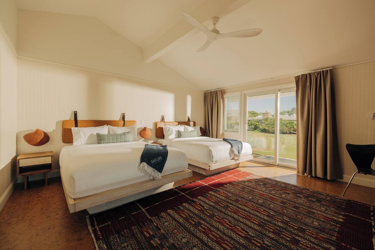A hotel room with two beds, a ceiling fan, a large window with curtains, and a view of greenery outside.