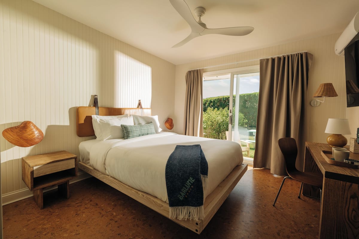 A hotel room with a bed, a desk, a chair, a nightstand, a ceiling fan, and a view of greenery through the window.