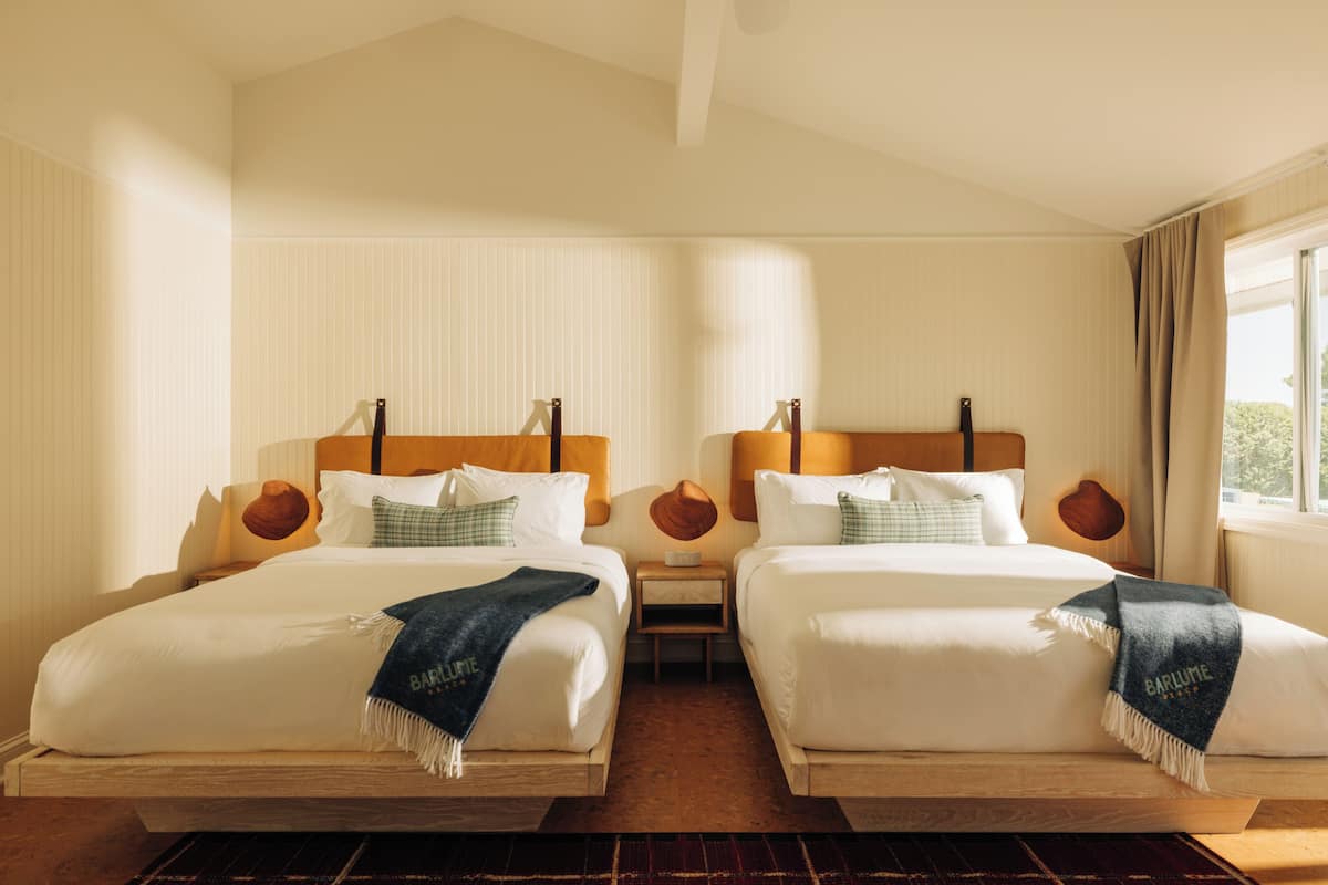 A hotel room with two beds, a wooden headboard, a small table, and a window with curtains.