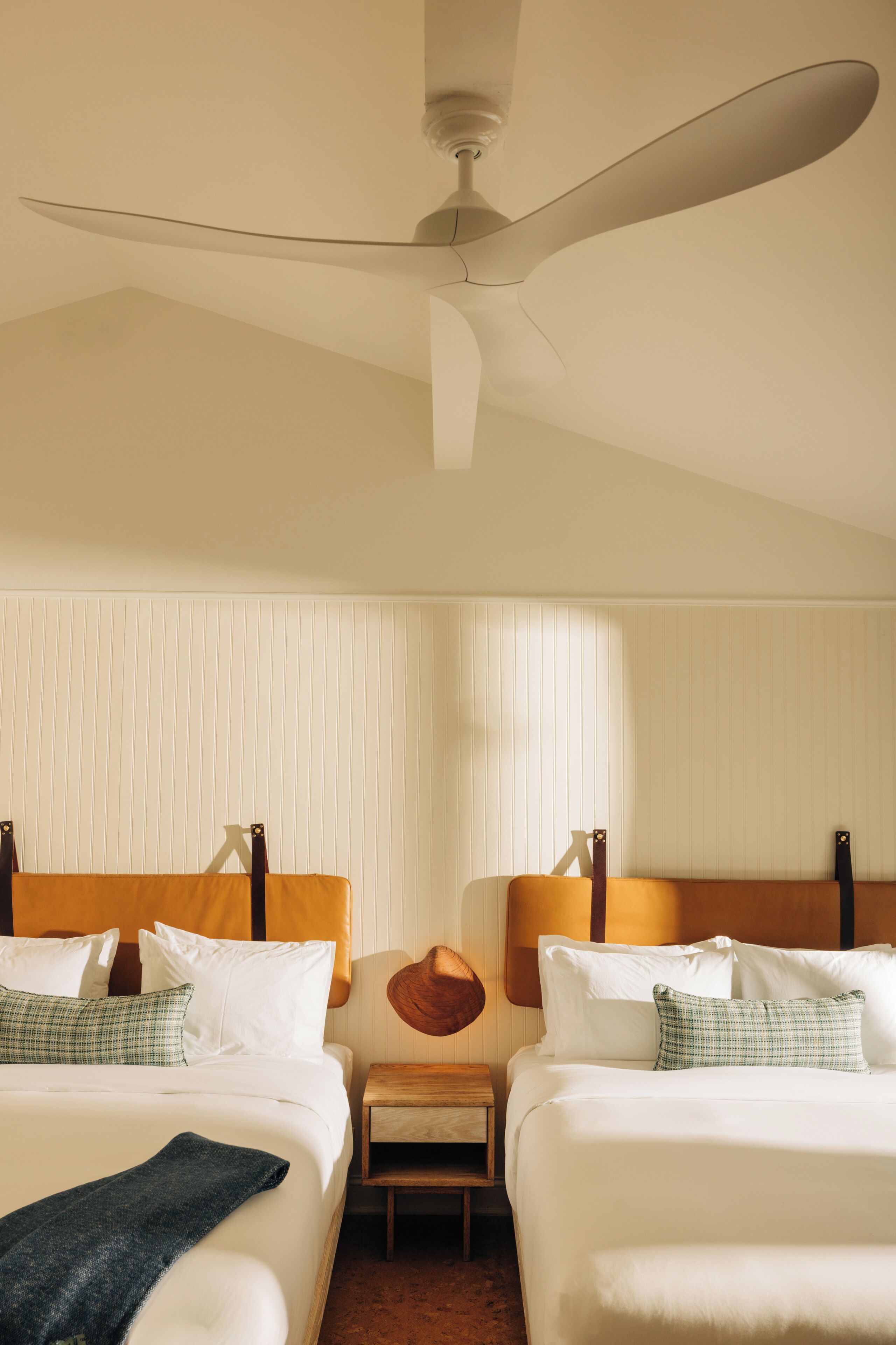 A hotel room with two beds, a ceiling fan, a small table, and a wall-mounted lamp.