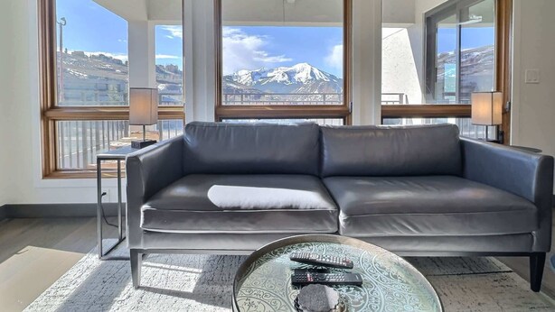 Un salon moderne avec un canapé en cuir gris, une table basse en verre dotée de télécommandes et de grandes fenêtres offrant une vue sur les montagnes.