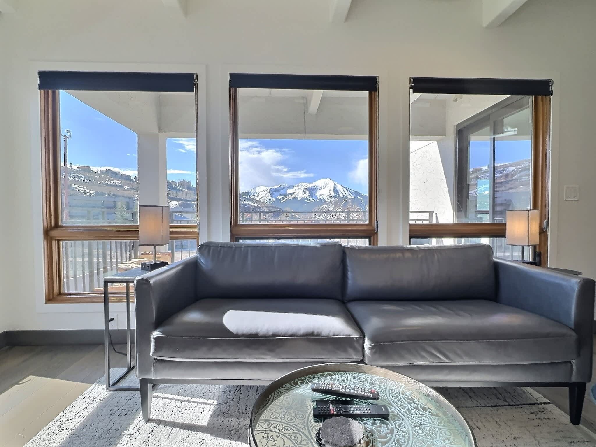 A modern living room with a grey leather sofa, a glass coffee table with remote controls, and large windows offering a mountain view.