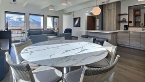 Une chambre d’hôtel moderne dotée d’une table ronde en marbre, de chaises en osier, d’un coin cuisine et de grandes fenêtres offrant une vue sur les montagnes.