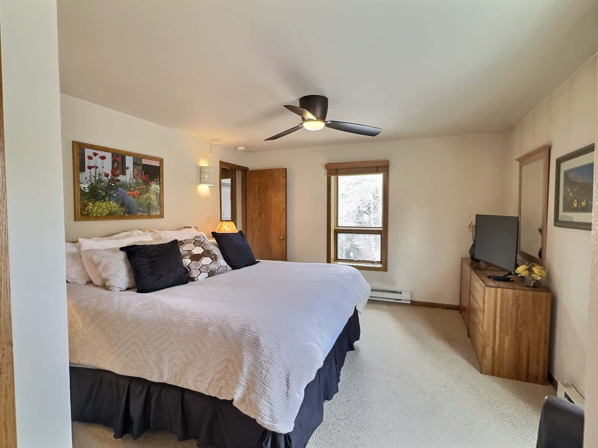 A bedroom with a bed, a ceiling fan, a TV on a wooden dresser, and a window.