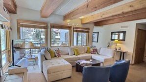 A spacious living room with a white sofa, blue armchair, and a coffee table. Large windows provide a view of the outside. - The Plaza 239 | Walk to CBMR Lifts | 2 Hot Tubs+Sauna | 3BR Sleeps 8 (Crested Butte)