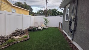A backyard with artificial grass, a white fence, a small table with chairs, and a tree. - Little oasis  2-bedroom house in fantastic St. Petersburg with AC (St. Petersburg)