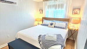 Un lit bien fait, recouvert d’une couvre-lit à motifs bleus et blancs, avec deux oreillers, une table de chevet dotée d’une lampe et un banc noir.