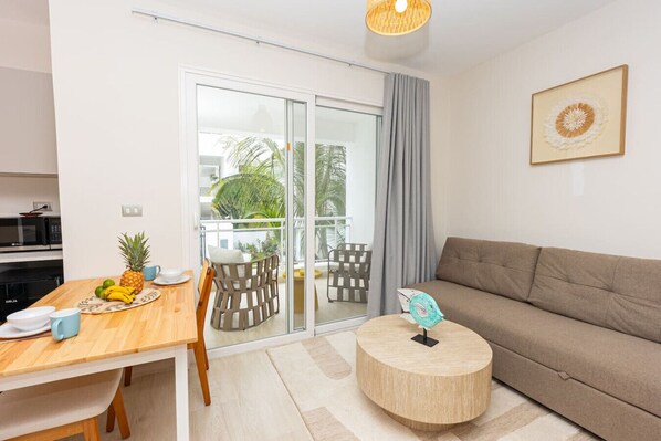A modern living room with a sofa, a dining table with chairs, a small round table with a decorative item, and a sliding glass door leading to a balcony with outdoor furniture.