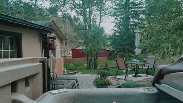 A hot tub with a person relaxing, a patio area with outdoor furniture, and a view of trees and a building in the background.