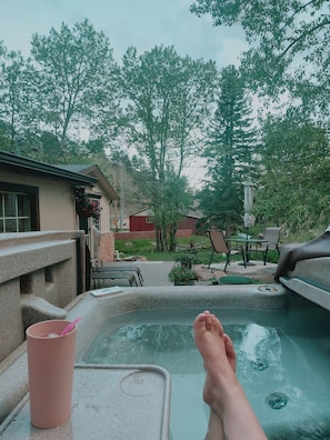 Ein Whirlpool mit einer entspannten Person, eine Terrasse mit Gartenmöbeln und im Hintergrund Bäume sowie ein Gebäude.
