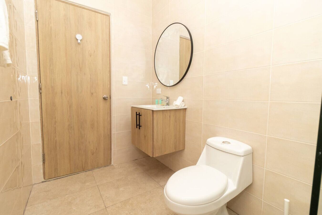 Une salle de bain avec une porte en bois, des toilettes, un lavabo avec un miroir et des murs recouverts de carreaux beiges.