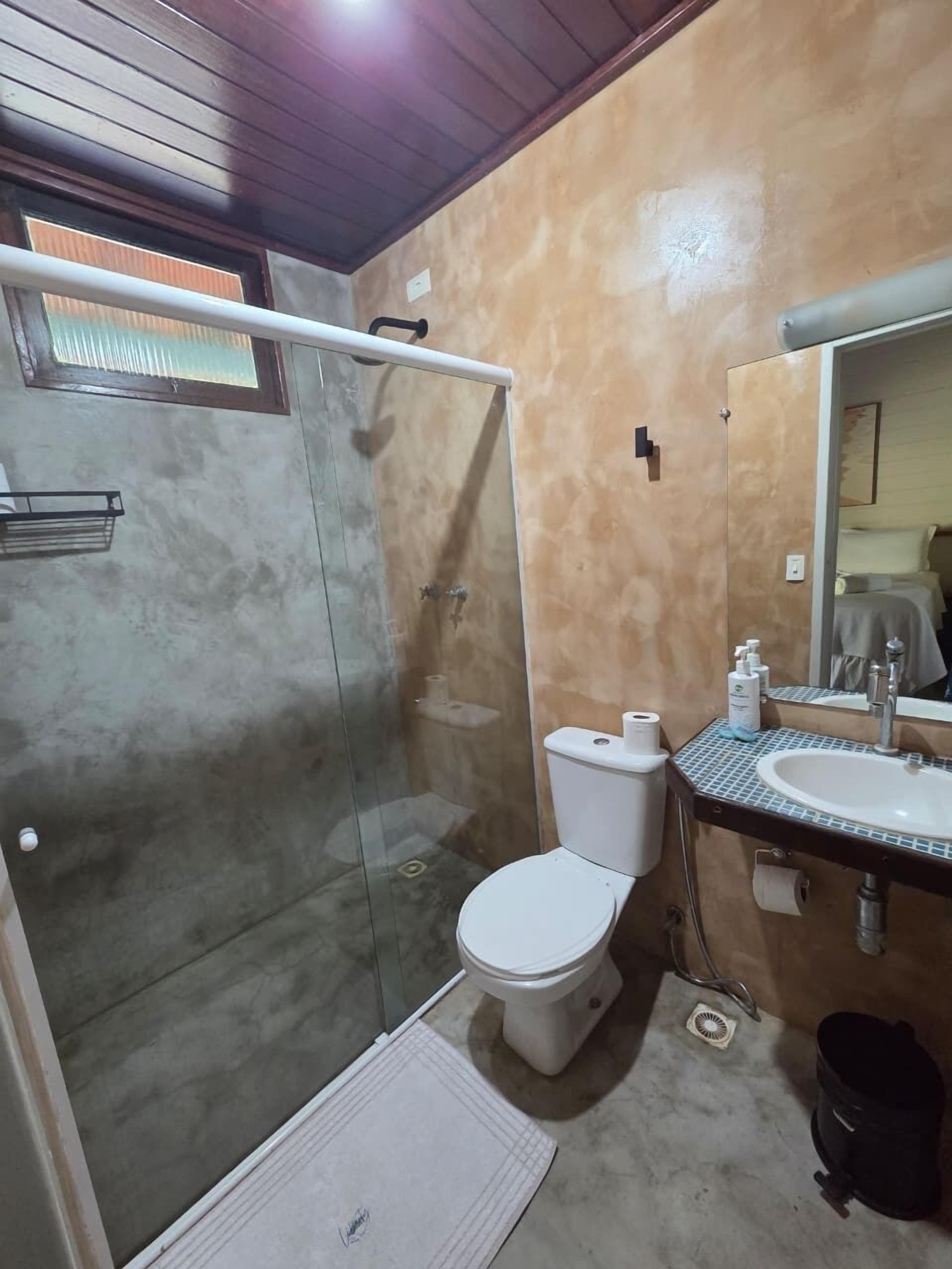 A bathroom with a glass-enclosed shower, a toilet, a sink with a mirror, and a small trash bin.