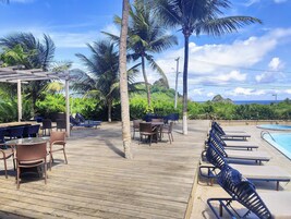 A wooden deck with lounge chairs and tables, a pool, and palm trees.