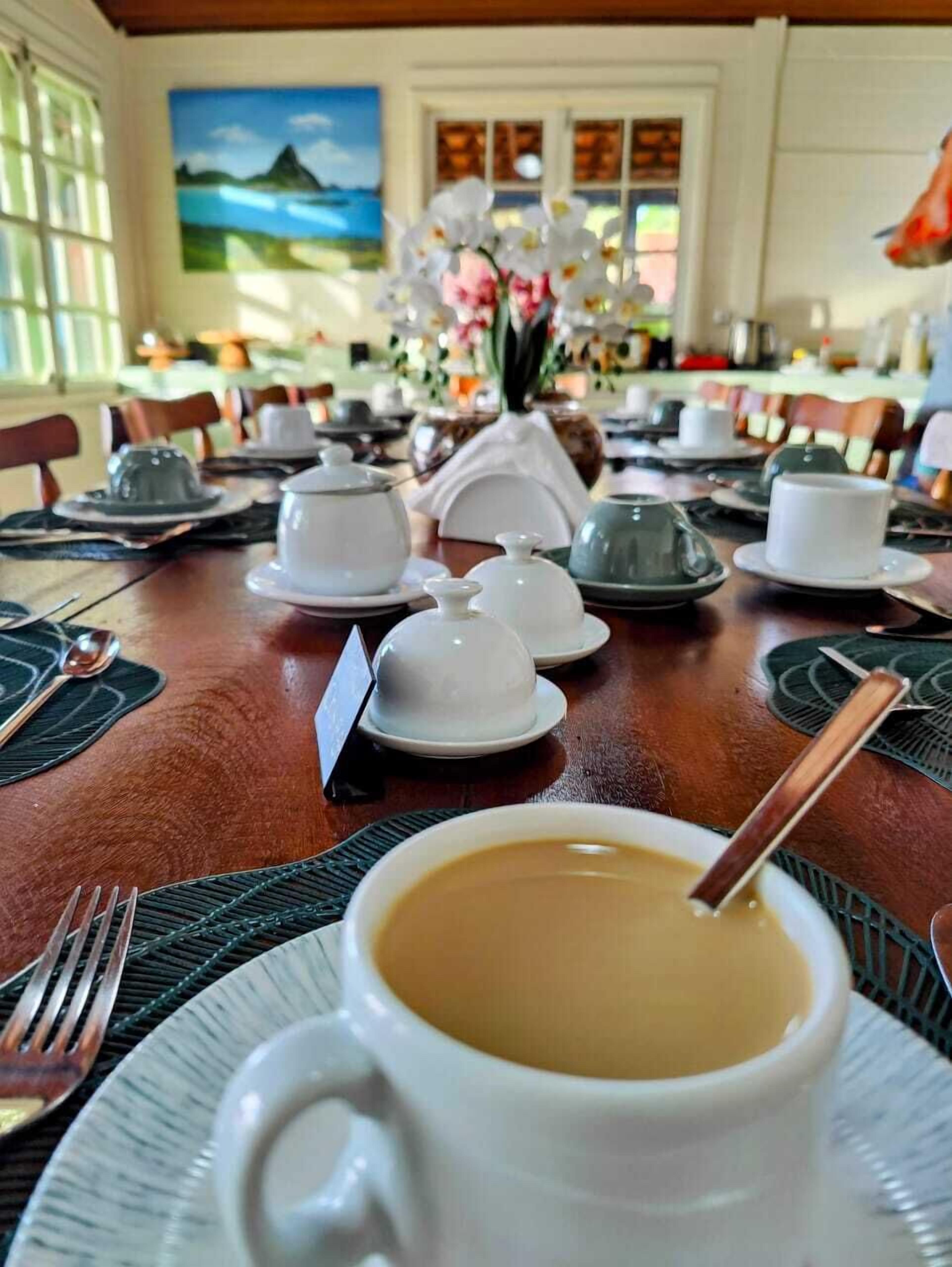 Une table à manger dressée avec des théières, des tasses et des soucoupes blanches, un vase rempli de fleurs et un tableau accroché au mur.