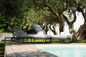 Un espace aménagé au bord de la piscine, avec des chaises longues et une table, entouré d’arbres et d’un bâtiment blanc.