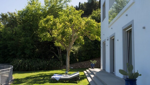 Un edificio moderno con facciata bianca, un giardino alberato e una zona prendisole con lettini.