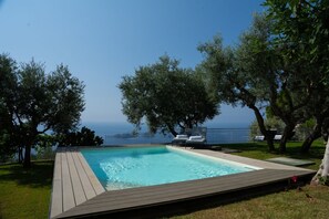 Une piscine rectangulaire avec une terrasse en bois, entourée d’arbres et offrant une vue sur la mer.
