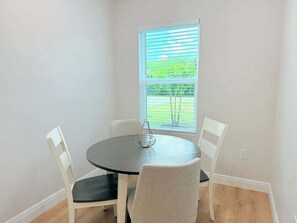 Un petit coin repas avec une table ronde et quatre chaises, une fenêtre avec des stores et une vue sur la verdure à l’extérieur.