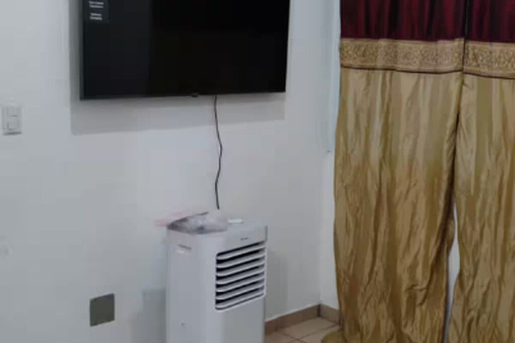 Huoneessa on seinälle asennettu televisio, lattialla seisova ilmastointilaite ja kuviollinen reunus, joka on ommeltu verhoon.