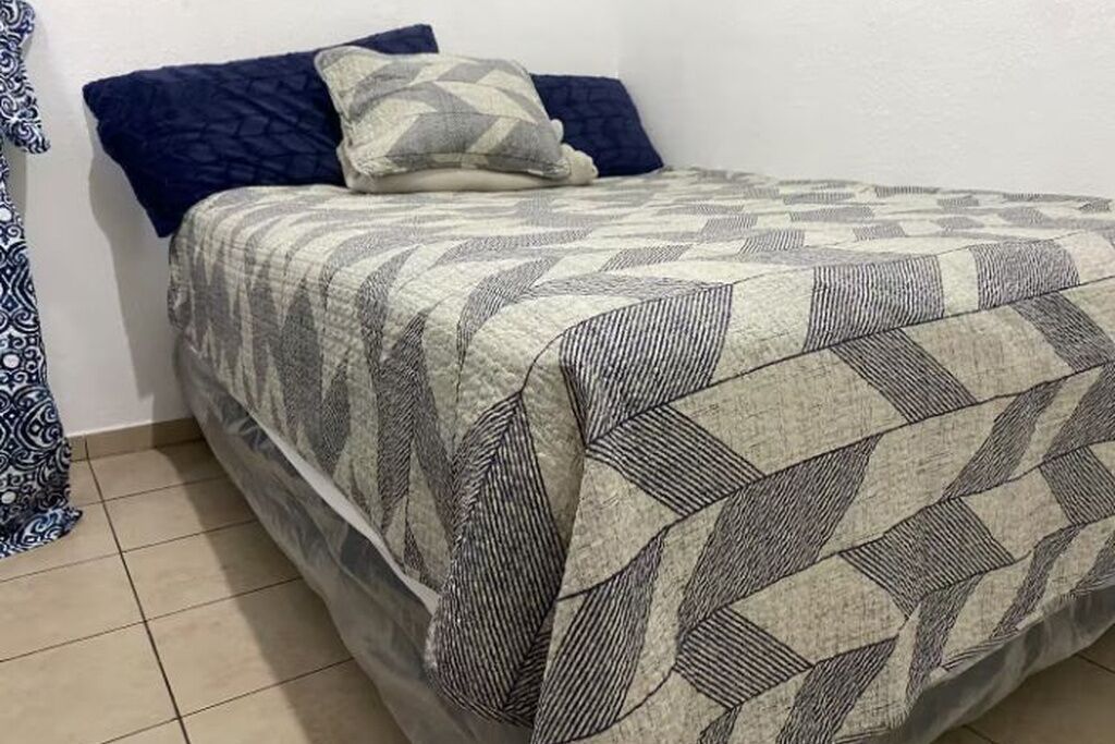 Ein Bett mit einem gemusterten Bettüberwurf, einem Kissen und einem blauen Kopfkissen auf einem Fliesenboden.