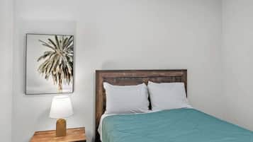 A single bed with a wooden headboard, a teal bedspread, and a white pillow. A nightstand with a lamp and a framed picture of a palm tree is to the left.