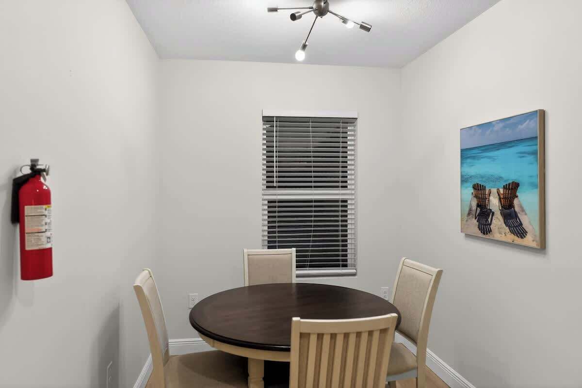 Una habitación con una mesa redonda, cuatro sillas, un ventilador de techo, un extintor, una ventana con persianas y un cuadro de una escena de playa.