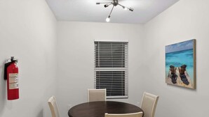 A room with a round table, four chairs, a ceiling fan, a fire extinguisher, a window with blinds, and a painting of a beach scene. - Relax in Englewood 3BR 2BA Home Close to Beaches (Englewood)