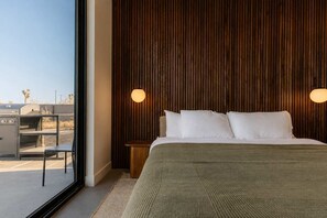 Ein Schlafzimmer mit großem Fenster, einem Bett mit weißer Bettwäsche, einem hölzernen Kopfteil und einem Nachttisch mit Lampe.