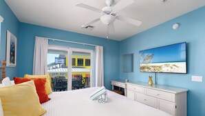 Quarto de hotel com cama, escrivaninha, cadeira, ventoinha no teto e um quadro na parede.