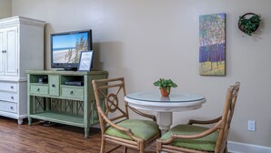Een kamer met een televisie op een kast, twee rieten stoelen met groene kussens, een klein tafeltje met een bloempot en twee aan de muur hangende schilderijen.