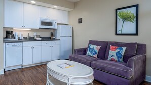 Een knus woongedeelte met een kitchenette, een paarse bank met decoratieve kussens, een rond rieten bijzettafeltje en een ingelijste botanische print aan de muur.