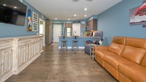 A living room with a brown leather sofa, a television mounted on the wall, and a kitchen area in the background. - Harbour House #212 "Shore Thing" (Fort Myers Beach)