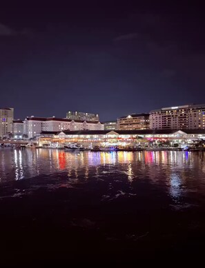 Un paisaje urbano frente al mar de noche, con edificios iluminados y reflejos en el agua.