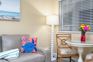 Ein Zimmer mit einer Couch, einem Sessel, einem Tisch mit einer Vase, einer Lampe und einem Fenster mit Jalousien.