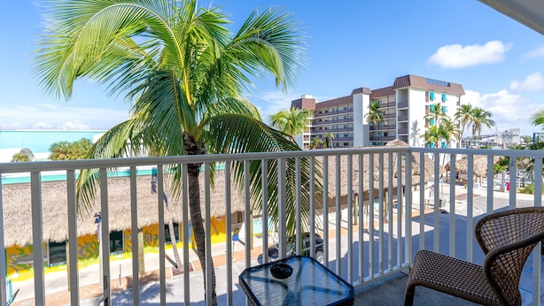 Een balkon met rieten stoelen, een kleine tafel en uitzicht op een strand met palmbomen en een gebouw op de achtergrond.