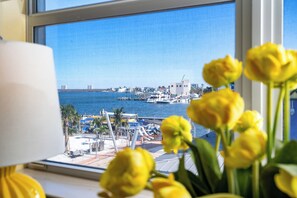 Uitzicht door een raam, met gele bloemen op de voorgrond, een jachthaven met boten, en gebouwen in de verte.