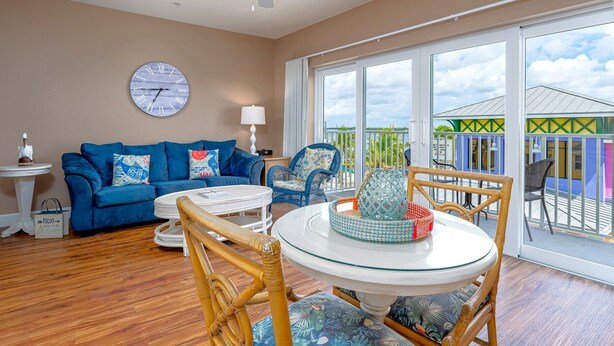 Un salon avec un canapé bleu, une table ronde en verre, des fauteuils en osier, une horloge et de grandes fenêtres donnant sur un bâtiment aux couleurs vives.