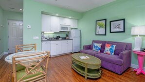 A compact living space with a kitchenette, a purple sofa with decorative pillows, a small round table, and a wicker ottoman. - Harbour House #310 "Sandpiper Retreat" (Fort Myers Beach)