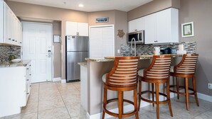 Una cocina con armarios blancos, un refrigerador de acero inoxidable, un microondas y un salpicadero de azulejos. Hay tres taburetes de bar de madera con asientos acolchados.