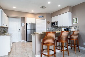 A kitchen with white cabinets, a stainless steel refrigerator, a microwave, and a tiled backsplash. There are three wooden bar stools with cushioned seats. - Harbour House #312 “Castaway Dreams” (Fort Myers Beach)