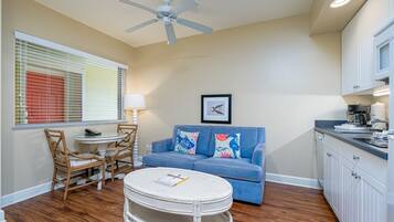 A compact living space with a dining area, a sofa, a ceiling fan, a kitchen area, and a window with blinds.